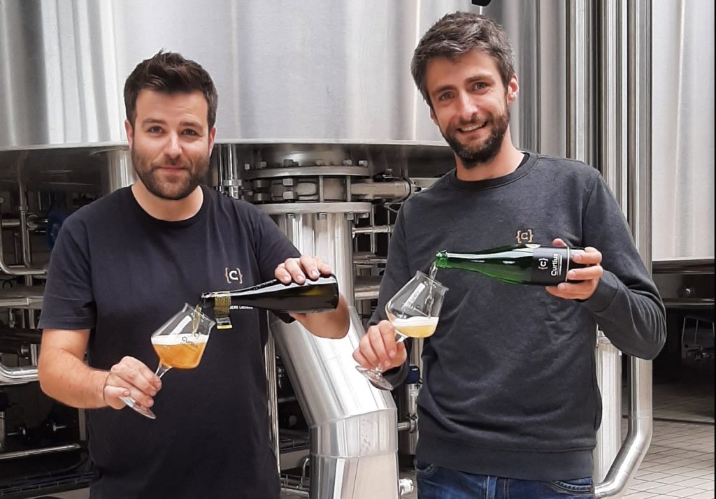 Zwei Männer in T-Shirts gießen Bier aus Flaschen in Gläser vor Braukesseln
