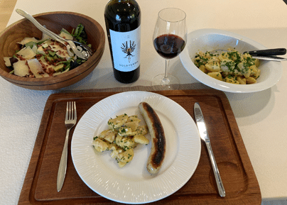 Table dressée avec assiette de saucisse et rösti, bouteille de vin rouge, verre de vin, salade et plat de pâtes