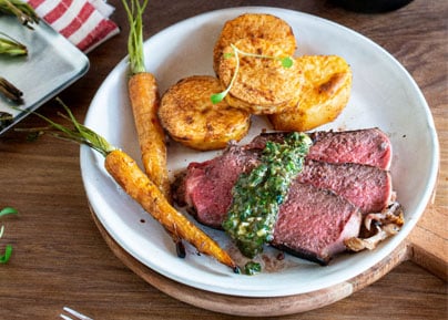 Assiette de viande rouge tranchée accompagnée de légumes rôtis et sauce verte sur planche de bois